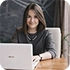 Woman with long dark hair smiling while using a silver laptop at a wooden table.
