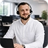 Smiling man with a beard sits at a desk with a laptop in a bright office.