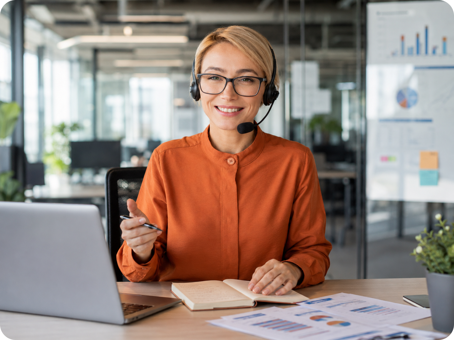 Untitled designнpng | Stream Telecom Customer service agent wearing a headset and glasses, smiling at a desk with a laptop, notebook, and charts in a bright office setting.